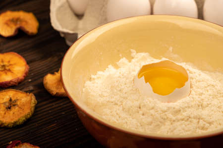 A broken white chicken egg in a bowl with wheat flour, dried apple chips and several whole eggs in a tray made of white cardboard, close-up selective focusの写真素材