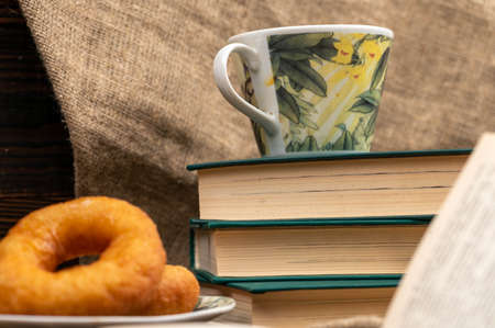 A cup of strong tea on a saucer, donuts on a plate and a stack of books on the table.の写真素材