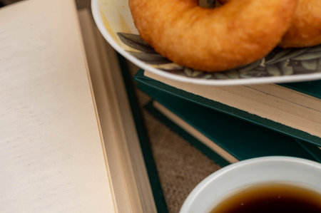 A cup of strong tea on a saucer, donuts on a plate and a stack of books on the table.