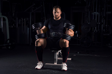 Impressive athlete sits on a bench in the gym with two dumbbells on his legsの写真素材