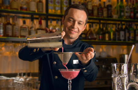 Charming bartender prepares a delicious cocktail. Strain the drink through a strainer and pour into a cocktail glassの写真素材