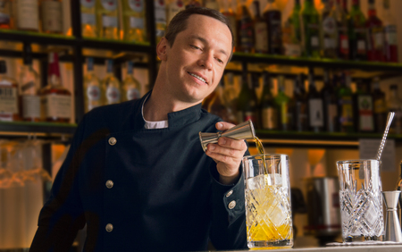 Charming bartender is pouring alcohol from a jigger into a mixing glass in order to cook an excellent drinkの写真素材