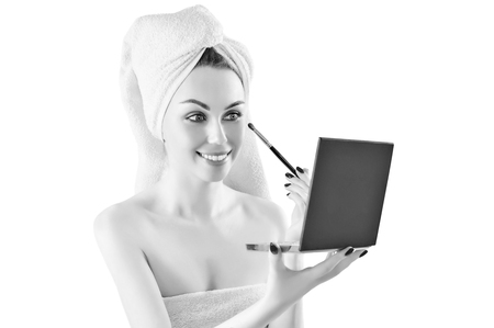 Delightful girl after a shower with a knotted towel on her head holds a set with makeup in her hands and paints her eyelids with a professional tassel. Mixed media. A concept for selling cosmetics.の写真素材