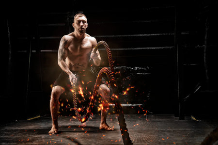 The athlete works with crossfit ropes in the gym. Flames, sparks and smoke. The concept of sports,...