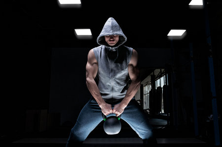 Athlete squat with kettlebell in the gym. Strong man in a sweatshirt with a hood. Crossfit,...