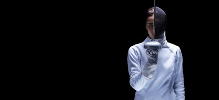 Charming girl in a fencer costume posing on a black background. The sword separates the head into a...