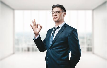 Real estate agent stands in a bright office and shows the ok sign to the camera. The concept of the sale of real estate, search for rental space. Mixed mediaの写真素材
