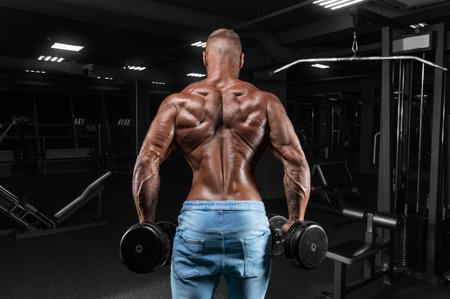The athlete does an exercise in the gym with dumbbells. Back view. Shrugs. Fitness, bodybuilding, powerlifting concept. Mixed mediaの写真素材
