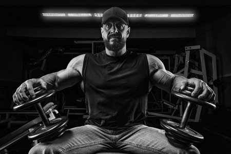 Muscular man sits on a bench with dumbbells in the gym. Fitness and bodybuilding concept. Mixed...