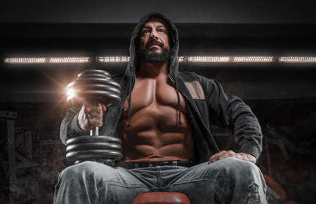 Muscular man sits on a bench with a dumbbell in the gym. Fitness and bodybuilding concept. Mixed mediaの写真素材