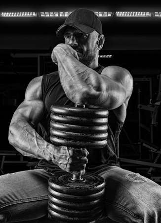 Muscular man sits on a bench with a dumbbell in the gym. Fitness and bodybuilding concept. Mixed mediaの写真素材