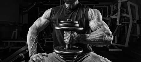 Muscular man sits on a bench with a dumbbell in the gym. Fitness and bodybuilding concept. Mixed mediaの写真素材