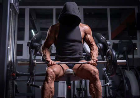 Noname image of a powerful athlete in a hoodie sitting on a bench in a gym. Fitness and bodybuilding concept. Mixed mediaの写真素材