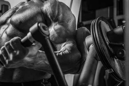 Black and white image of a powerful athlete sitting on a bench in a gym and pumping biceps. Fitness and bodybuilding concept. Mixed mediaの写真素材