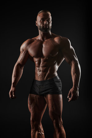 Muscular man shows his muscles against the background of a black wall. Bodybuilder, male naked torso, abs. Mixed mediaの写真素材