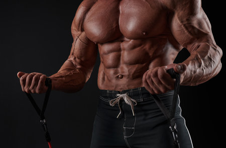 Muscular man working out with elastic band, isolated on black wall background. Bodybuilder, male naked torso, abs. Mixed media. Close up photoの写真素材