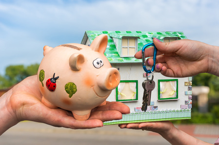 Real estate agent giving house keys to a new property owner, who is paying all saving money from piggy bank on blurred backgroundの写真素材