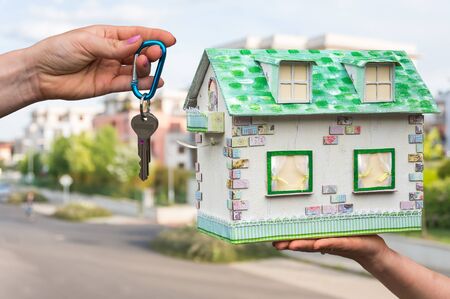 Real estate agent giving house keys to a new property owner, who is holding model house from paper on blurred backgroundの写真素材