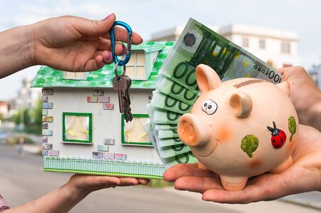 Real estate agent giving house key and model house to a new property owner, who is paying by euro money from piggy bank on blurred backgroundの写真素材
