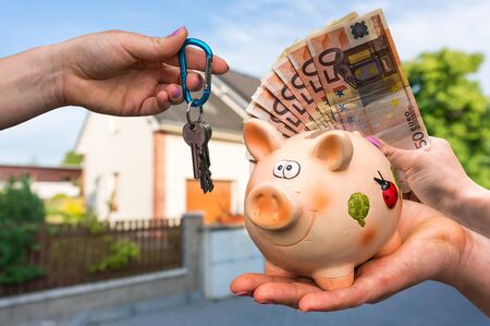 Real estate agent giving house keys to a new property owner, who is paying by euro money from piggy bank on blurred backgroundの写真素材
