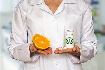 Young doctor holding two halves of fresh orange and bottle of pills with vitamin C and compare themの写真素材