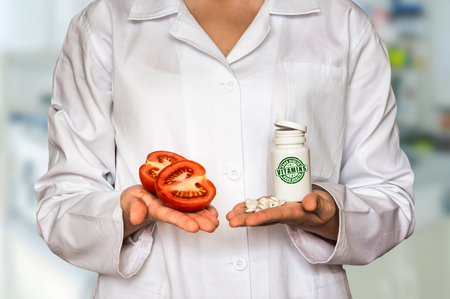 Young doctor holding fresh tomato and bottle of pills with vitamins and compare themの写真素材