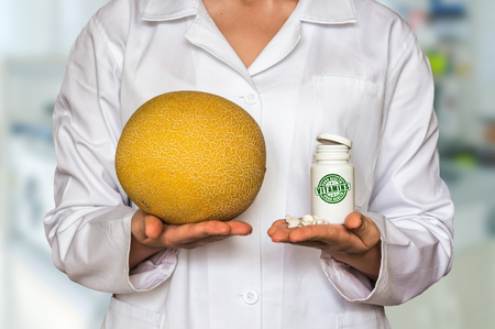 Young doctor holding fresh yellow melon and bottle of pills with vitamins and compare themの写真素材