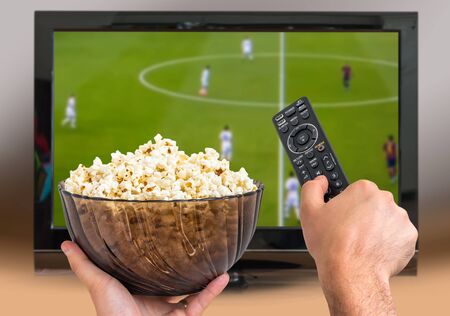 Man is watching football match on TV and holds remote controller and popcorn in handsの写真素材