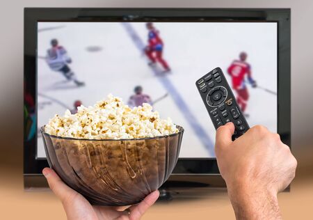 Man is watching hockey match on TV and holds remote controller and popcorn in handsの写真素材