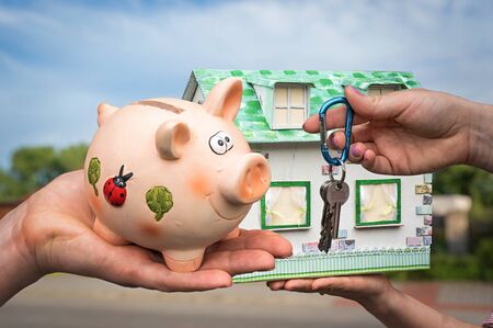 Real estate agent giving house key and model house to a new property owner, who is paying last saving money from piggy bank on blurred backgroundの写真素材