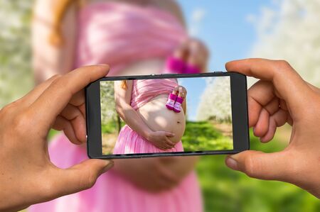 Woman hands with mobile cell phone to take a photo of belly of a pregnant girl and small booties in the blooming fruit gardenの写真素材