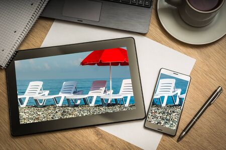 Digital tablet on business table with sunbathing beds and red umbrella on screen and mobile phone, paper, pen, laptop and cup of coffee - above viewの写真素材