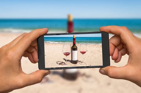 Woman hands with mobile cell phone to take a photo of bottle of wine and glasses on the beachの写真素材