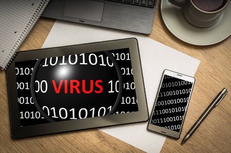 Digital tablet on business table with binary code with virus on screen and mobile phone, paper, pen, laptop and cup of coffee - above viewの写真素材