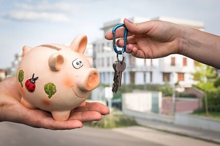 Real estate agent giving flat keys to a new property owner, who is paying last saving money from piggy bank on blurred backgroundの写真素材