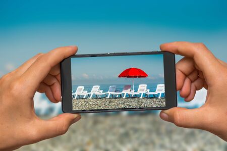 Woman hands with mobile cell phone to take a photo of sunbathing plastic beds and red umbrella on the beachの写真素材