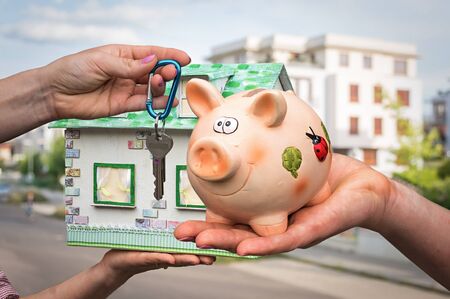 Real estate agent giving flat keys to a new property owner, who is paying last saving money from piggy bank on blurred backgroundの写真素材