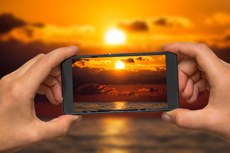 Woman hands with mobile cell phone to take a photo of sunset and seaの写真素材