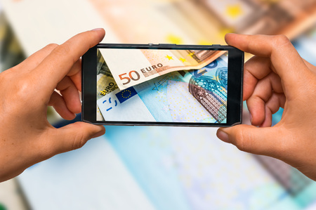 Woman hands with mobile cell phone to take a photo of euro money banknotesの写真素材