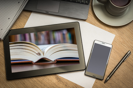 Digital tablet on business table with open book in library on screen and mobile phone, paper, pen, laptop and cup of coffee - above viewの写真素材