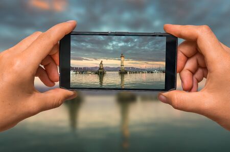 Woman hands with mobile cell phone to take a photo of evening seascape with lighthouse in harborの写真素材