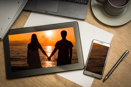 Digital tablet on business table with silhouette of a loving couple at sunset on screen and mobile phone, paper, pen, laptop and cup of coffee - above viewの写真素材