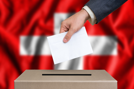 Election in Switzerland. The hand of man putting his vote in the ballot box. Swiss flag on background.の写真素材