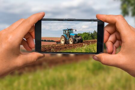 Woman hands with mobile cell phone to take a photo of tractor at work on a fieldの写真素材