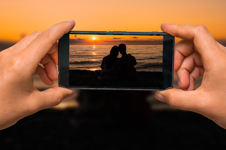 Woman hands with mobile cell phone to take a photo of silhouette of a loving couple at sunset near seaの写真素材