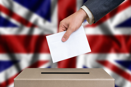 Election in United Kingdom. The hand of man putting his vote in the ballot box. United Kingdom flag on background.の写真素材