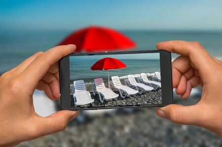 Woman hands with mobile cell phone to take a photo of sunbathing plastic beds and red umbrella on the beachの写真素材