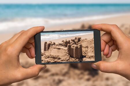 Woman hands with mobile cell phone to take a photo of sand castle on the sea shoreの写真素材