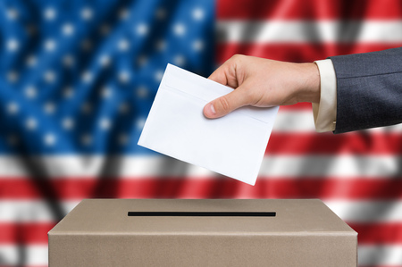 Presidential election in United States of America. The hand of man putting his vote in the ballot box. American flag on background.の写真素材