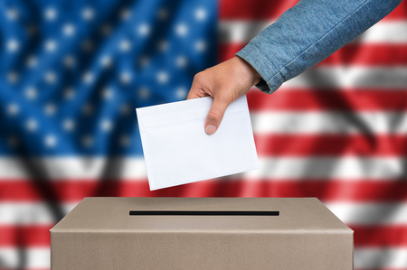 Election in United States of America. The hand of woman putting her vote in the ballot box. American flag on background.の写真素材